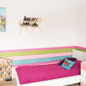 May include: A white wooden horse head shelf with multiple miniature horses on the shelves. The shelf is mounted on a white wall with a pink, green, and blue striped border. A white bed with a pink bedspread is in the foreground.