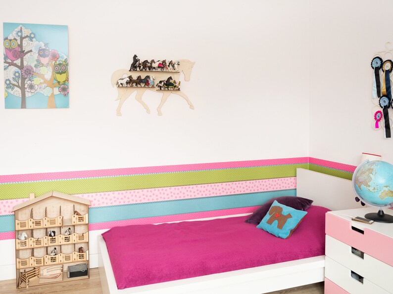 May include: A white wooden horse head shelf with multiple miniature horses on the shelves. The shelf is mounted on a white wall with a pink, green, and blue striped border. A white bed with a pink bedspread is in the foreground.