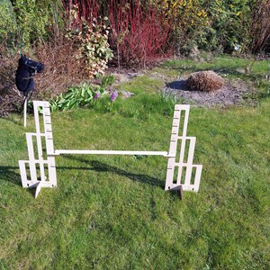 Puede incluir: Un recorrido de obst&aacute;culos de madera para un caballo de juguete, que consta de dos vallas de madera blancas conectadas por una tabla de madera. El recorrido est&aacute; instalado en un c&eacute;sped.