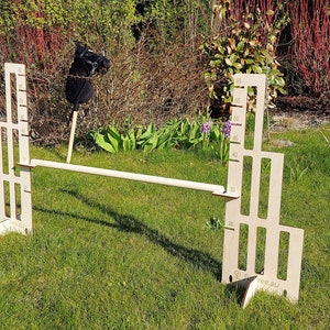 Puede incluir: Un obst&aacute;culo de salto de altura ajustable de madera para caballos, con una barra de madera blanca y una escala de altura marcada en cent&iacute;metros. El obst&aacute;culo est&aacute; instalado en un c&eacute;sped.