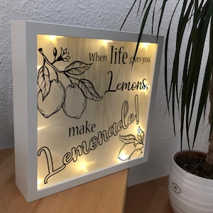 May include: White framed light-up sign with a black and white illustration of lemons on a branch. The sign reads "When life gives you lemons, make lemonade!"