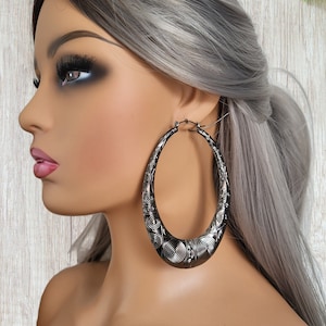 May include: Large silver hoop earrings with a detailed swirl pattern. The earrings are oval shaped and have a shiny finish.