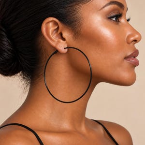 May include: A large, thin, circular hoop earring in a matte black finish. The earring features a simple, continuous wire design that forms a wide, open circle, providing a clean and understated look that rests against the side of the face.