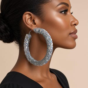 May include: A large hoop earring covered in iridescent, light-reflecting sequins that shimmer with shades of silver, blue, and purple. The circular earring features a thick, textured surface created by the dense application of small, round, metallic-finish sequins.