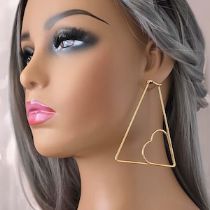 May include: Gold triangle hoop earrings with a heart cutout. The earrings are large and dangle from the earlobe.