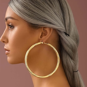 May include: A profile view showcases a person wearing large, gold hoop earrings. The earrings are circular with a ridged texture. The person has gray hair styled in a braid. The background is a solid, muted pink.