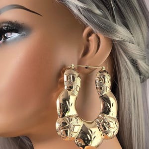 May include: Large gold hoop earrings with a textured, geometric design. The earrings are thick and have a rounded shape.
