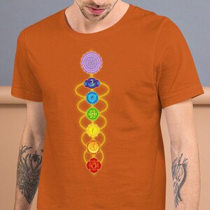 May include: A man is wearing an orange t-shirt with a colorful chakra symbol design. The symbol is a vertical line with seven circles, each a different color, representing the seven chakras.