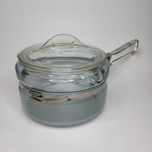 Può includere: Una casseruola vintage in vetro Pyrex con coperchio e manico in vetro trasparente. La pentola è di colore grigio-azzurro chiaro con una fascia metallica color argento intorno al centro. Il coperchio ha un manico ad anello.