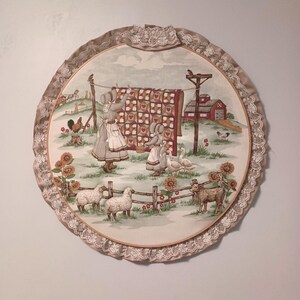 Puede incluir: Un tapiz circular de tela con una escena pastoral de dos figuras colgando una colcha en un tendedero. La imagen incluye un granero rojo, ovejas, gansos, un gallo y girasoles, todo enmarcado por una tela beige con un borde de encaje blanco.