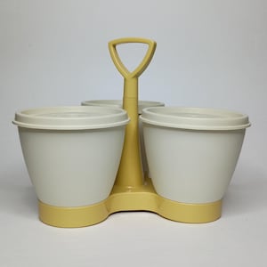 May include: A vintage yellow and white condiment caddy with a handle. The caddy has a triangular handle and holds three white containers with lids. The base and handle are yellow, and the containers are white.