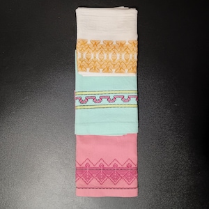 Può includere: Un set di tre strofinacci rettangolari piegati e impilati verticalmente. Lo strofinaccio superiore è bianco con un bordo geometrico dorato. Quello centrale è verde menta chiaro con un motivo a zigzag rosa e giallo. Quello inferiore è rosa con un bordo geometrico rosa scuro.
