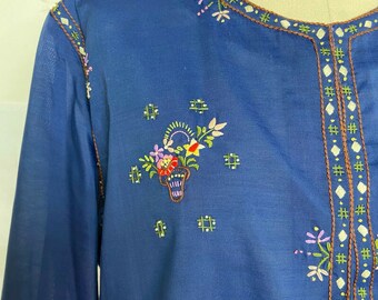 Puede incluir: Una túnica azul bordada con un diseño floral y ribete marrón. El bordado presenta una cesta de flores en tonos rojos, rosas, verdes y amarillos.