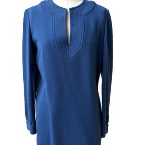 May include: A long-sleeved blue tunic top featuring a split V-neckline with decorative vertical stitching around the collar and down the front placket. The garment has a straight hemline and fitted cuffs, presented on a mannequin torso.