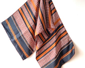 Può includere: Una sciarpa di seta con motivo a pied-de-poule e a righe nei toni del marrone, arancione e blu navy. La sciarpa è appesa su uno sfondo bianco. La sciarpa ha un'etichetta nell'angolo.