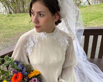 Op de afbeelding: Een vintage ivoorkleurige trouwjurk met een hoge halslijn en lange klokmouwen. De jurk heeft delicate witte bloemenapplicaties op de schouders en een lange, vloeiende sluier. Een kleurrijk boeket bloemen wordt vastgehouden.