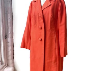 May include: A vibrant orange coat with a classic design, displayed on a mannequin. The coat features a notched collar, long sleeves, and three large buttons down the front. The tailored silhouette suggests a vintage or retro style.