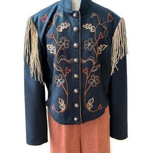 May include: A dark teal jacket featuring a high collar, silver-toned metal buttons, and long beige fringe along the shoulders. The front is decorated with intricate floral embroidery in shades of blue, red, and gold, paired with solid orange trousers.