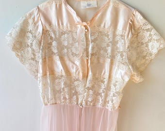 May include: A vintage peach-colored nightgown with lace detailing and a sheer, flowing skirt. The top features a delicate lace overlay on the bodice and sleeves, with a satin-like fabric underneath. The gown is buttoned down the front and has a small bow at the neckline.