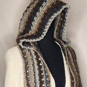 Peut inclure: Une écharpe à capuche en crochet de couleur marron, beige et gris avec un motif texturé. L'écharpe est drapé sur un pull blanc.