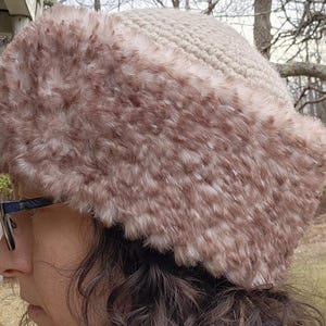 May include: A beige crocheted hat with a plush, faux fur trim in shades of brown and white. The hat is designed to keep the wearer warm and stylish during the colder months. The hat is a winter accessory.