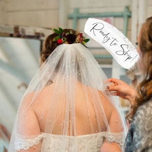 May include: A bride in a white lace wedding dress and veil with pearl accents. Another person is adjusting the veil. The bride wears a floral hairpiece. The text "Ready to Ship!" is visible.