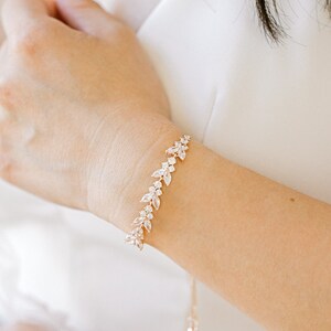 May include: Rose gold bracelet featuring a leaf design, embellished with clear crystals. A matching rose gold necklace with a teardrop pendant is also visible. The jewellery is worn with a white dress.