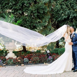 May include: A couple embraces outdoors on their wedding day. The bride wears a white wedding dress with a long train and veil. The groom is in a navy blue suit. The veil is billowing in the wind, creating a dramatic effect.