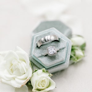 May include: A wedding ring set featuring a silver band and a square-cut diamond engagement ring, displayed in a light blue velvet ring box. White roses and greenery accent the scene, creating a romantic and elegant composition.
