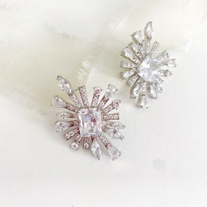 May include: A pair of silver earrings with a starburst design. Each earring has a large, rectangular, clear gemstone at the centre, surrounded by smaller, clear, teardrop and round-cut gemstones.