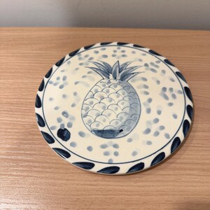 May include: Round ceramic coaster with a hand-painted pineapple design. The coaster features a cream-colored background with blue polka dots and a blue pineapple in the center. The edge is decorated with a blue leaf pattern.