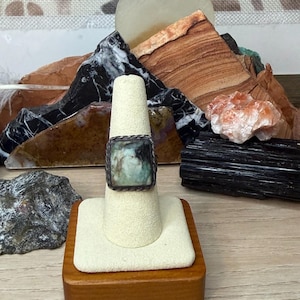 May include: A square ring with a light green stone set in a dark metal frame. The ring is displayed on a white ring holder with a wooden base. The background features various rocks and minerals.