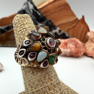 May include: A gold-toned ring featuring an array of colorful gemstones. The ring is adorned with various stones in shades of red, brown, green, and white. The ring is displayed on a textured, tan ring holder, with a backdrop of natural rock formations.
