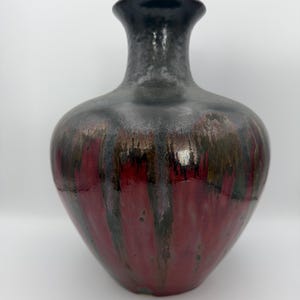 May include: A ceramic vase with a glossy finish. The vase has a dark gray neck and a body that transitions from dark red to black and green. The vase has a wide base and a narrow neck, suitable for displaying flowers or as a decorative piece.