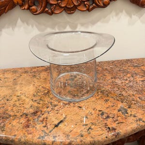 May include: A clear glass vase shaped like a top hat with a wide, flat brim and a cylindrical base. The glass is transparent and smooth, featuring a distinct wide rim that flares outward from the top of the main vessel, creating a unique silhouette.