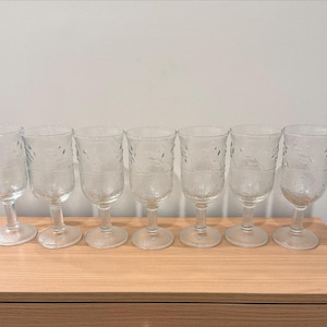 May include: Set of six clear, stemmed wine glasses. Each glass features a textured floral pattern and a rounded bowl. The glasses are arranged on a light-colored wooden surface against a plain white wall.