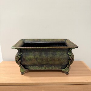 May include: A rectangular, antique-style planter with a distressed green and brown finish. The planter has decorative leaf accents on the sides and feet. The interior is black, and the planter sits on a wooden surface.