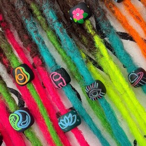 May include: A set of brown synthetic dreadlocks with colorful accents. The dreadlocks are decorated with black beads and colorful charms, including a flower, an avocado, a yin yang symbol, a lotus flower, and an eye. The dreadlocks are arranged in a fan shape on a white surface with a black background.