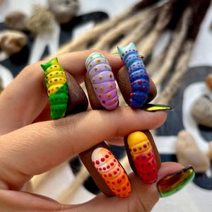 Puede incluir: Una mano muestra cinco diseños de uñas con temática de orugas. Las uñas presentan gradientes vibrantes de verde, amarillo, morado, azul y rojo, con patrones de puntos y rayas. Las uñas están pintadas sobre una base marrón oscuro.