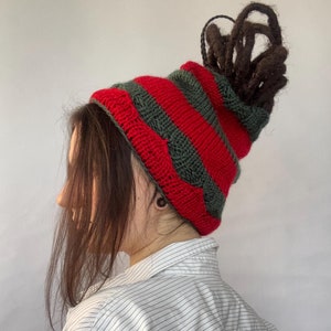 May include: A red and green striped knit beanie with a unique design. The beanie is worn on a person with long brown dreadlocks.