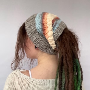 May include: A grey knitted hat with a colourful stripe pattern and a ponytail hole. The hat is worn by a person with long brown and green dreadlocks.