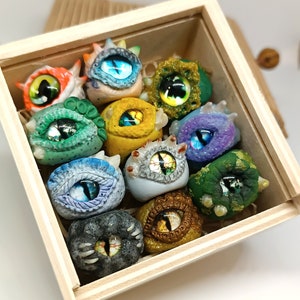Könnte beinhalten: Eine Holzkiste gefüllt mit 10 bunten, handgefertigten Polymer Clay Augen. Jedes Auge hat ein anderes Design und eine andere Farbe, darunter Grün, Blau, Lila und Gold. Die Augen sind perfekt, um jedem Bastelprojekt eine einzigartige Note zu verleihen.