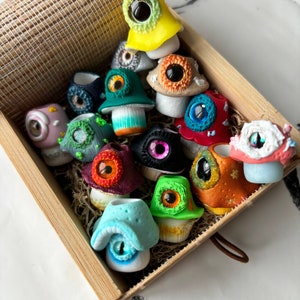 Puede incluir: Una caja de madera llena de figuritas de hongos hechas a mano y coloridas. Cada hongo tiene un ojo grande y saltones y un diseño único. Los hongos están hechos de arcilla polimérica y están pintados en varios colores, incluyendo rojo, verde, azul, amarillo y morado.