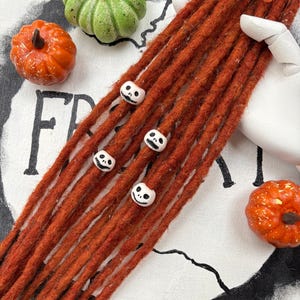 Op de afbeelding: Roestkleurige dreadlocks versierd met witte schedel kralen, gerangschikt met decoratieve pompoenen. Een groene pompoen en twee oranje pompoenen zijn rond de dreadlocks geplaatst. Het woord "FR" is zichtbaar op de achtergrond.