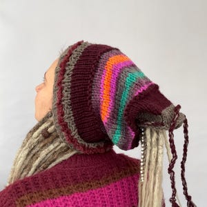 May include: A knitted hat with a striped pattern in shades of burgundy, gray, orange, pink, and teal. The hat has attached faux dreadlocks and tassels. The person is wearing a burgundy sweater with pink and brown stripes.