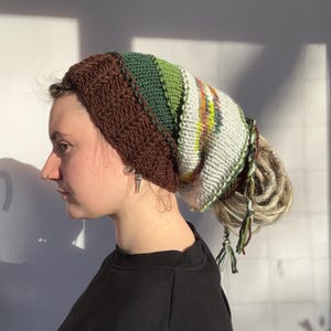 May include: A handmade, colourful beanie with a brown, textured band and a striped top in shades of green, white, and yellow. The beanie has attached dreadlocks with green and white tassels. The hat is designed for headwear.