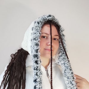 Könnte beinhalten: Eine weiße Kapuzenjacke mit schwarzem und weißem Pelzbesatz. Die Kapuze ist hochgezogen und die Jacke ist zugezippt. Die Person, die die Jacke trägt, hat lange braune Haare in Zöpfen.