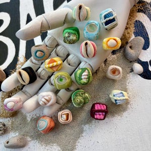 May include: A collection of colorful, handcrafted rings, each featuring unique seashell designs. The rings are displayed on a white hand, with a sandy texture in the background. The rings showcase a variety of colors and patterns, perfect for beach-themed jewelry.