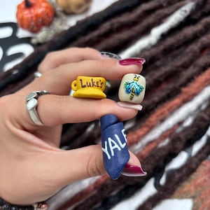 May include: Three small decorative charms made of clay. One charm is a yellow coffee mug with the text Luke's in brown. Another is a beige square with a blue and green dragonfly illustration. The third is a dark blue cylinder with the text YALE in white.