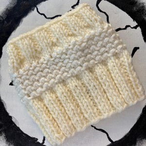 May include: A white knitted hat with a textured pattern. The hat has a band of white yarn with a slightly different texture.